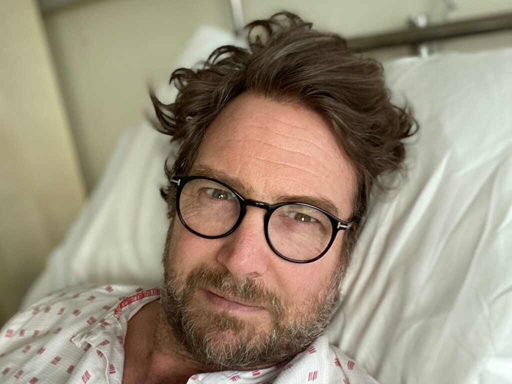 En mann med briller og skjegg ligger i en sykehusseng, iført en mønstret sykehuskjole. Sykehusveggen og medisinsk utstyr er synlig i bakgrunnen.