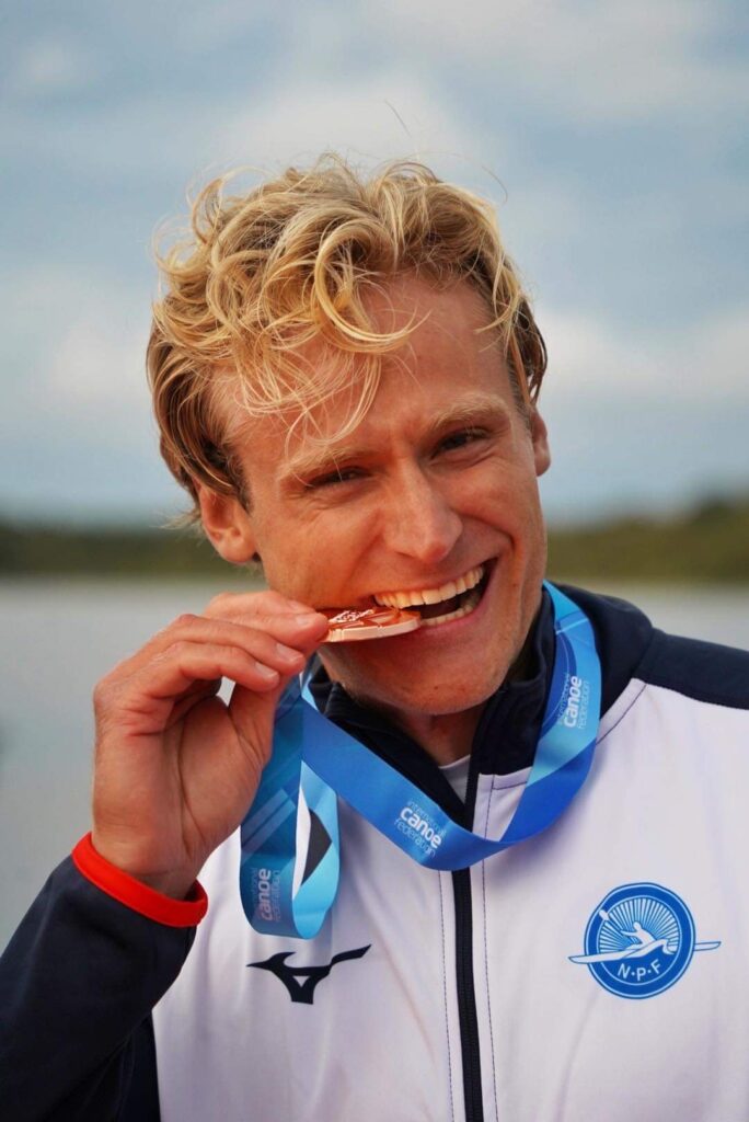 En mann iført sportsjakke og blått bånd med medalje biter på medaljen mens han smiler utendørs.