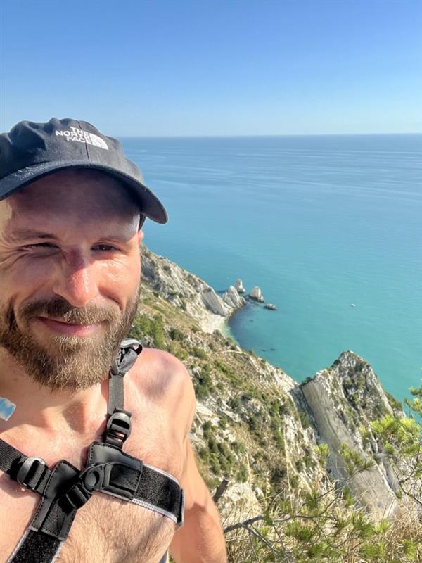 En mann iført lue og sele smiler til kameraet med en steinete kystlinje og blått hav i bakgrunnen under en klar himmel.