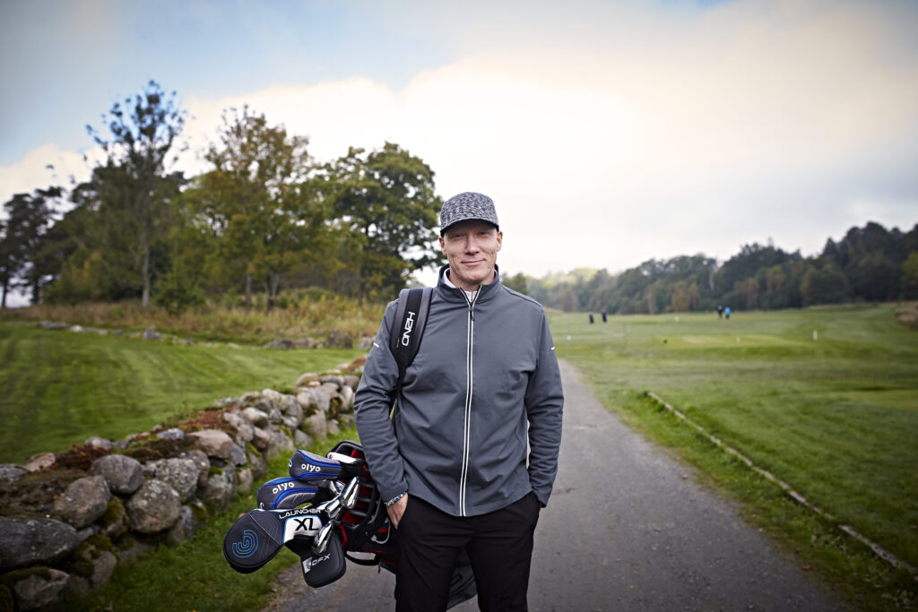 En person står på en golfbane med en golfbag i hånden, iført grå jakke og caps, med trær og gressplener i bakgrunnen.