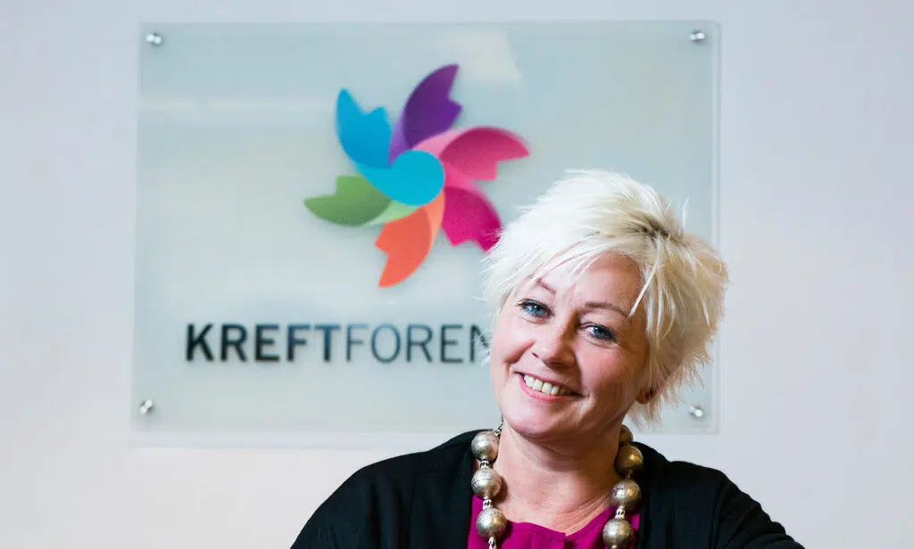 En person med kort lyst hår smiler foran et skilt med en fargerik logo og teksten «KREFTFOREN.