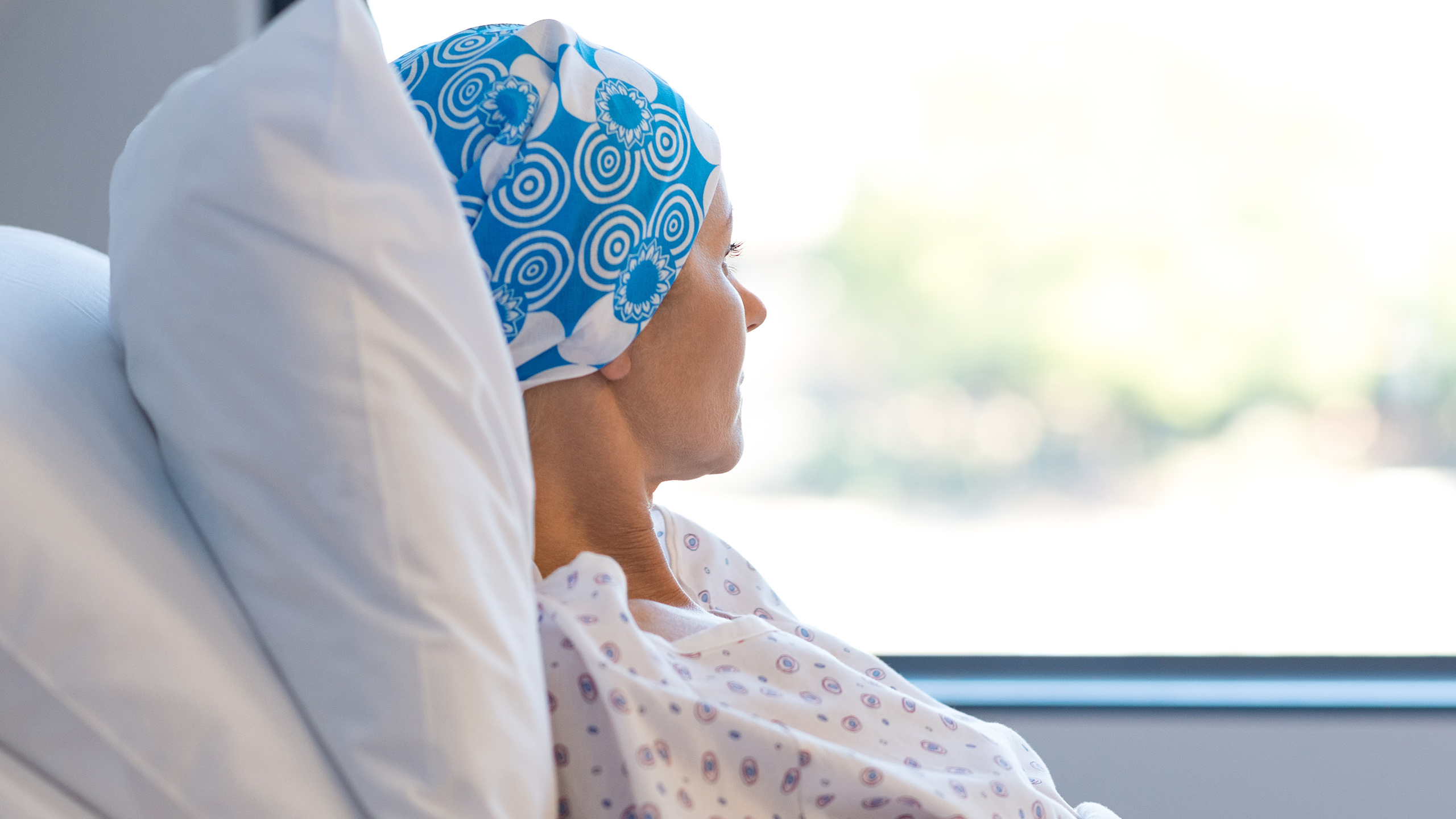 Illustrasjonsfoto av kreftpasient med blått tørkle på hodet, sittende i en sykeseng mens hun ser ut av vinduet.