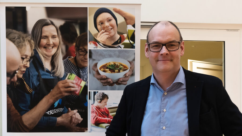 Tidligere kreftpasient foran en plakat med bilder fra Vardesenteret. Foto.