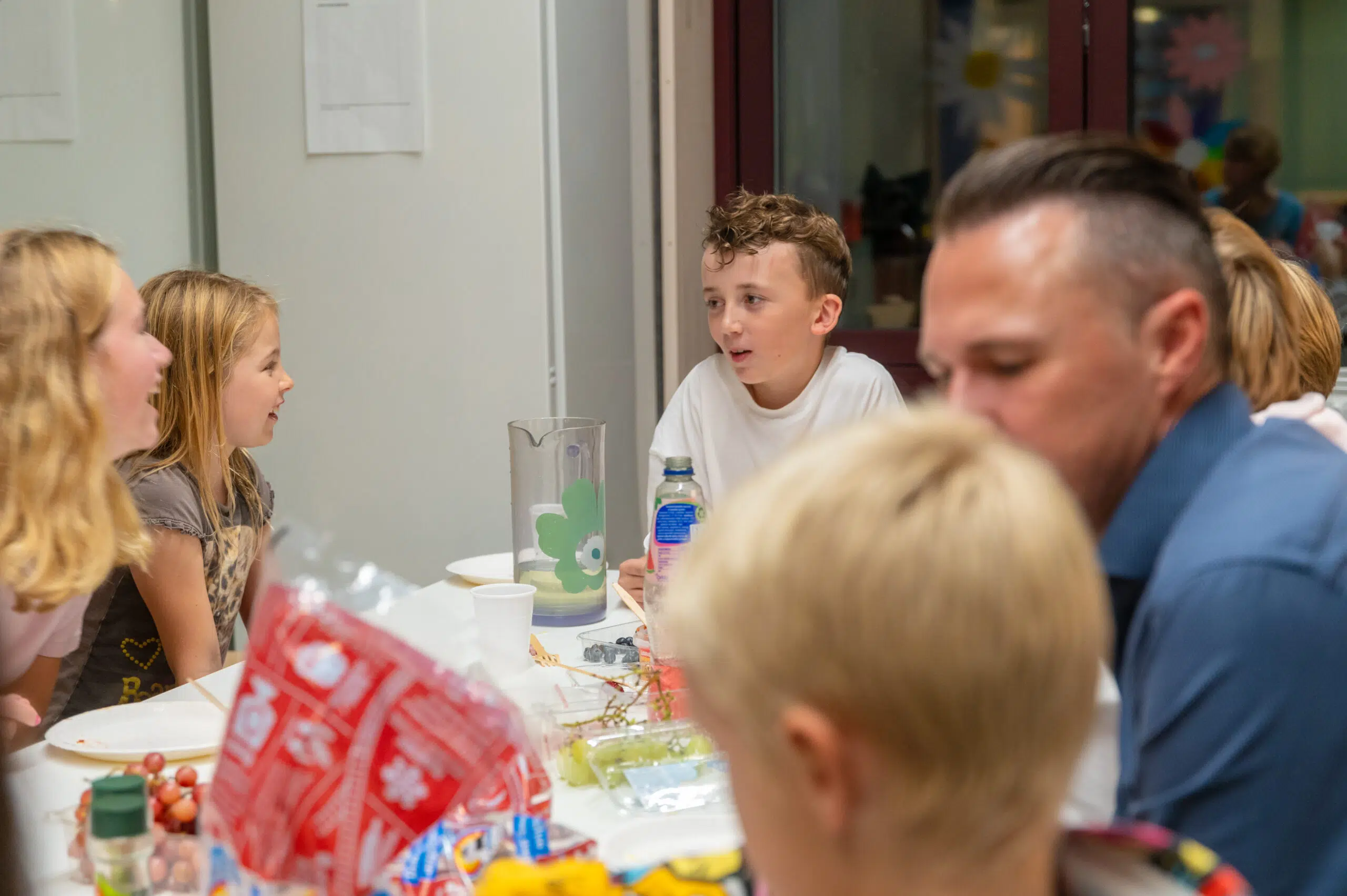 Fem personer, blant dem barn og voksne, sitter rundt et bord med snacks, drikke og mat, og snakker sammen i et godt opplyst rom.