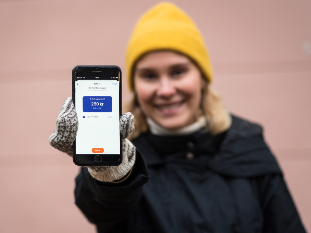 Kvinne med gul lue holder mobilen frem og viser appen Vipps