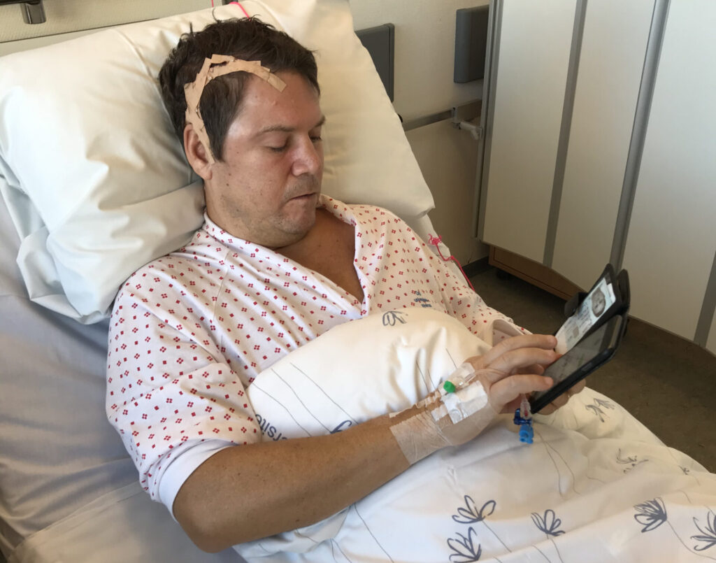 en mann som ligger i en sykehusseng og holder en mobiltelefon.