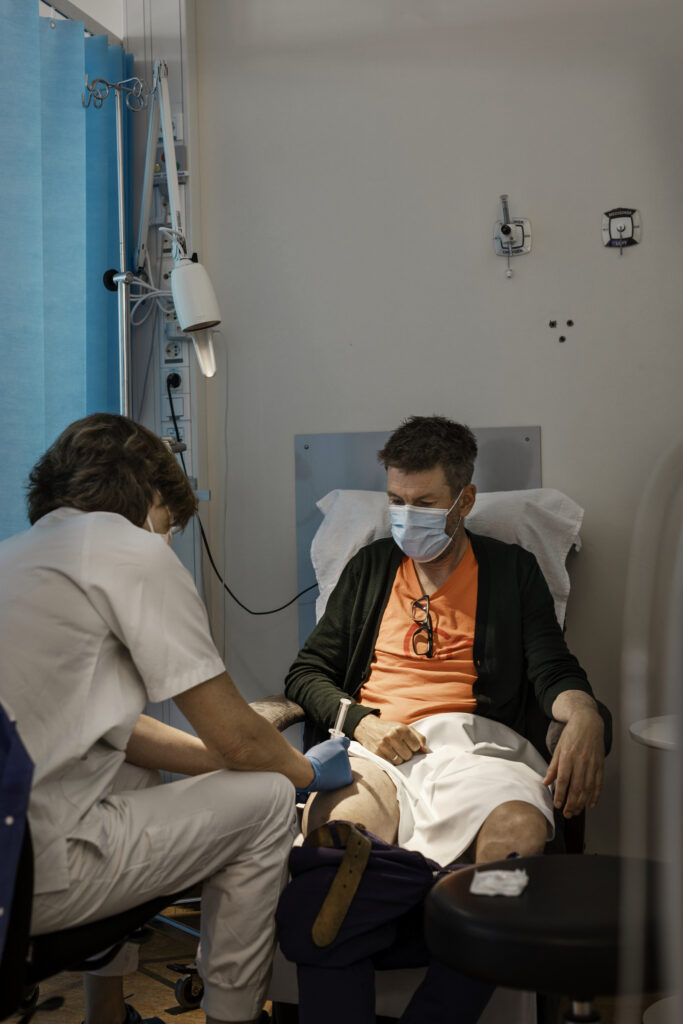 en mann som sitter i en sykehusseng med en annen mann iført ansiktsmaske.