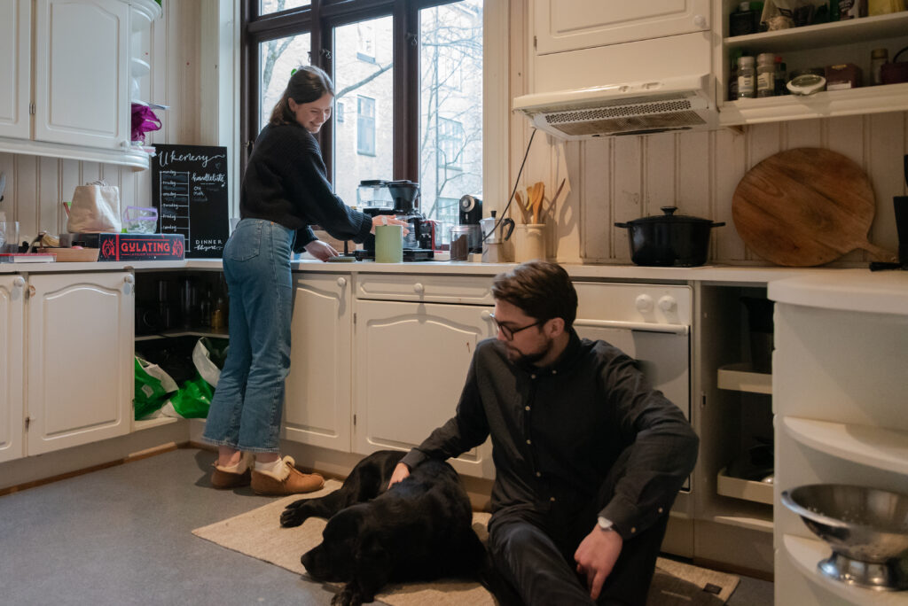 Adrian sitter på gulvet med en hund. Emilie lager kaffe.