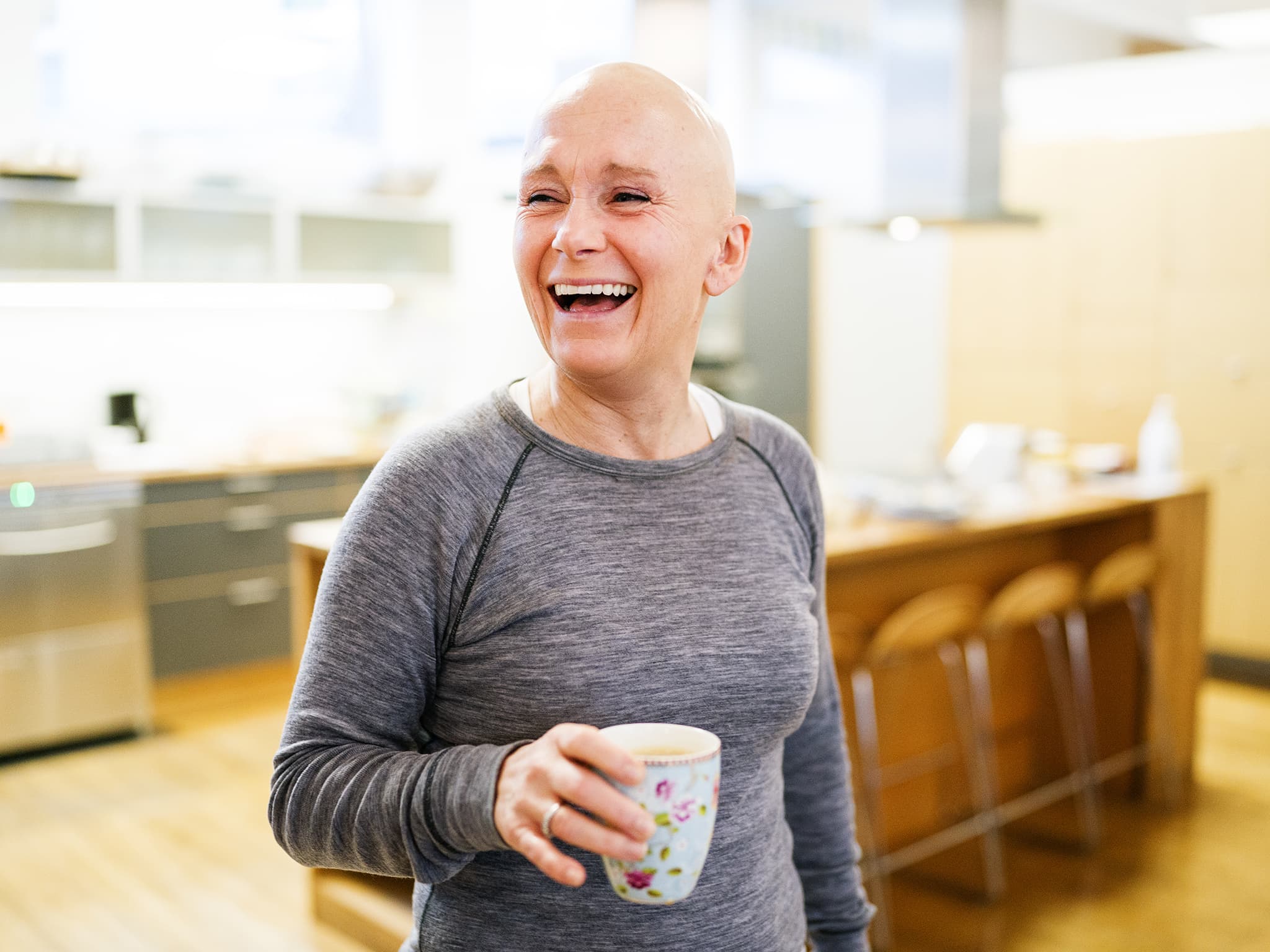 Kvinne uten hår på hodet holder en kaffekopp og smiler. Foto.