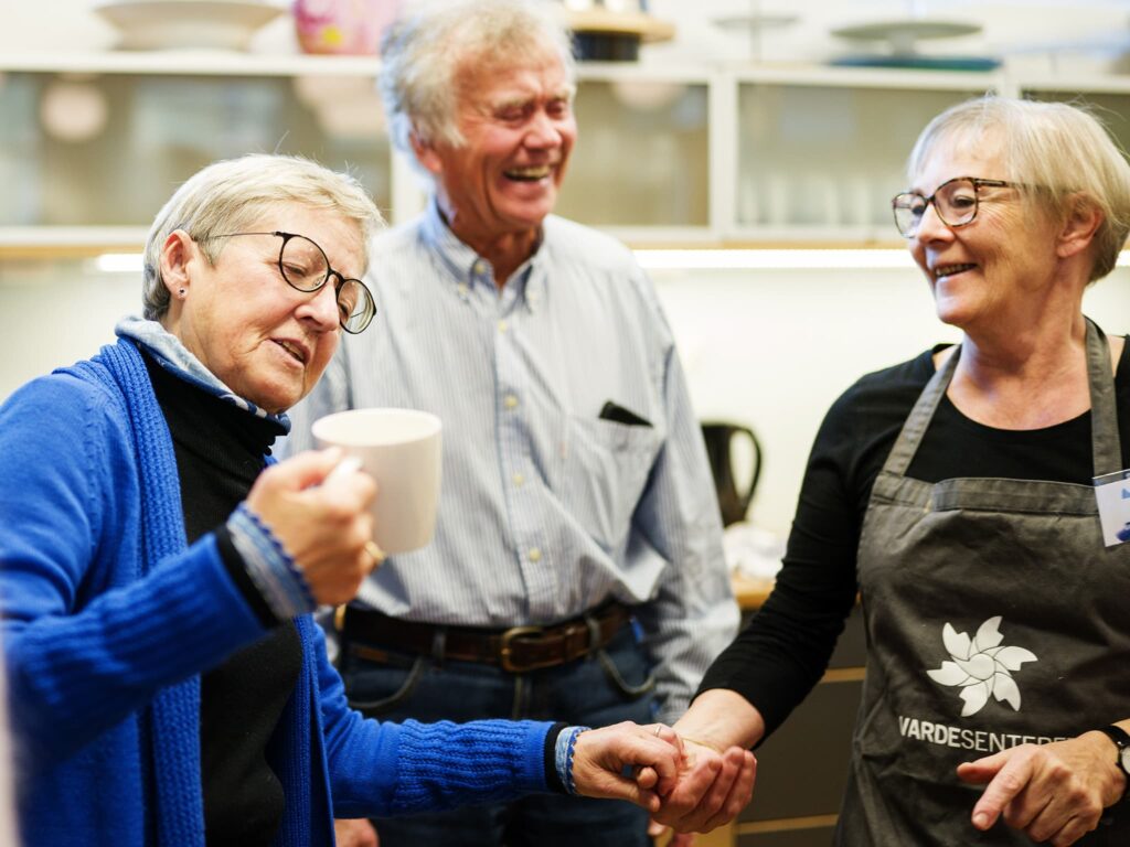 To eldre kvinner holder hender. Den ene kvinnen ser på en kaffekopp. I bakgrunnen står en mann og smiler. Foto.