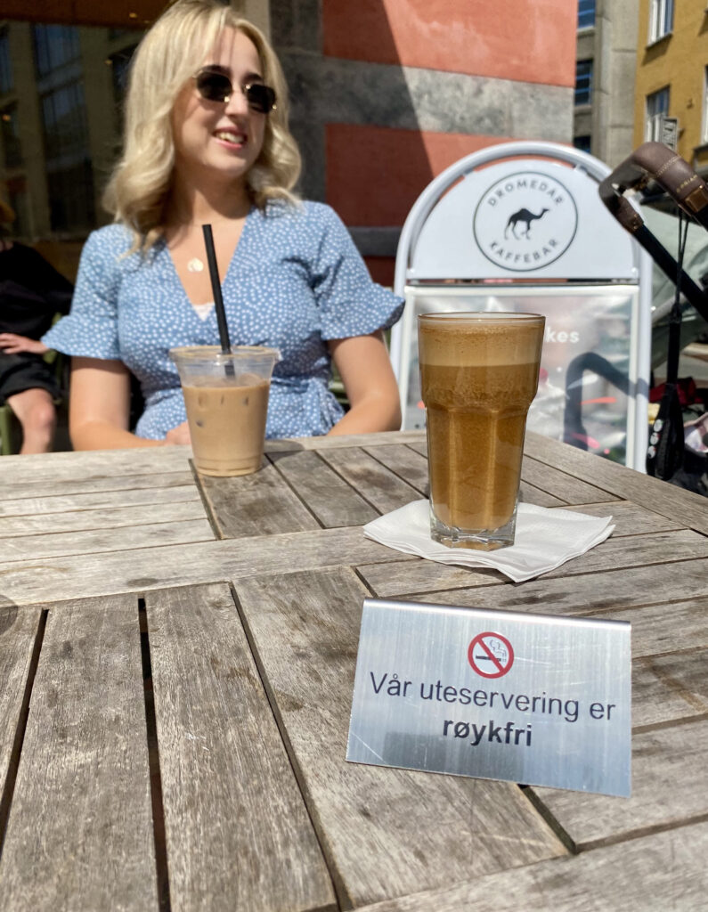 Kvinne drikker kaffe på kafe som har uteservering. Fremst i bildet er et et skilt med teksten "Vår uteservering er røykfri".