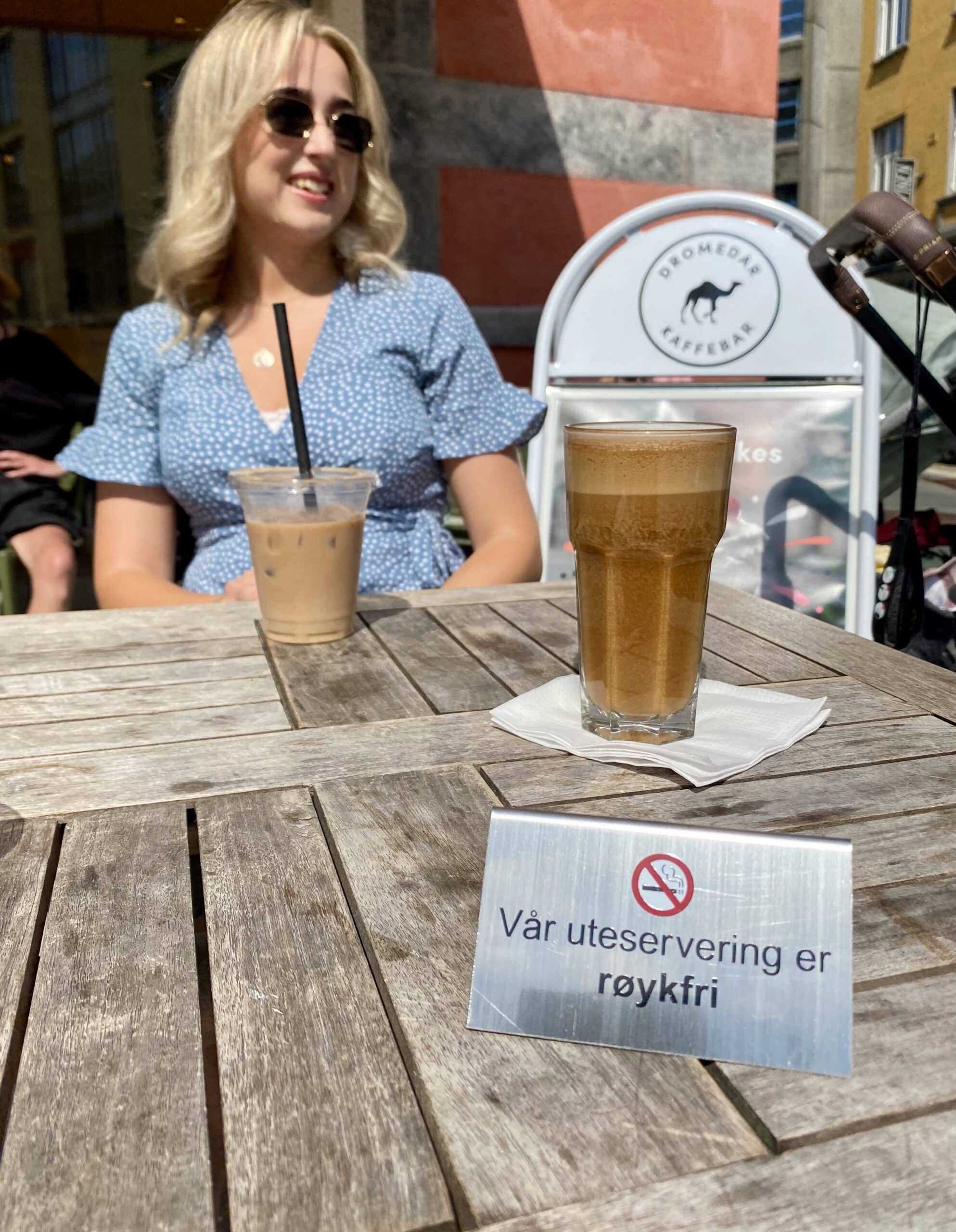 Kvinne drikker kaffe på kafe som har uteservering. Fremst i bildet er et et skilt med teksten Vår uteservering er røykfri.