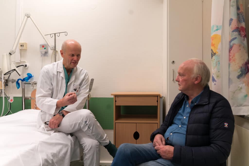 En mann som sitter i en sykehusseng og snakker med lege Viktor Berge.