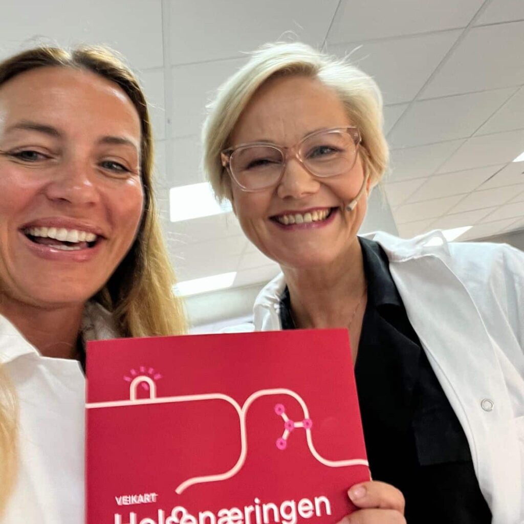 To kvinner i hvite laboratoriefrakker holder opp en bok med ordet helsingingen på.