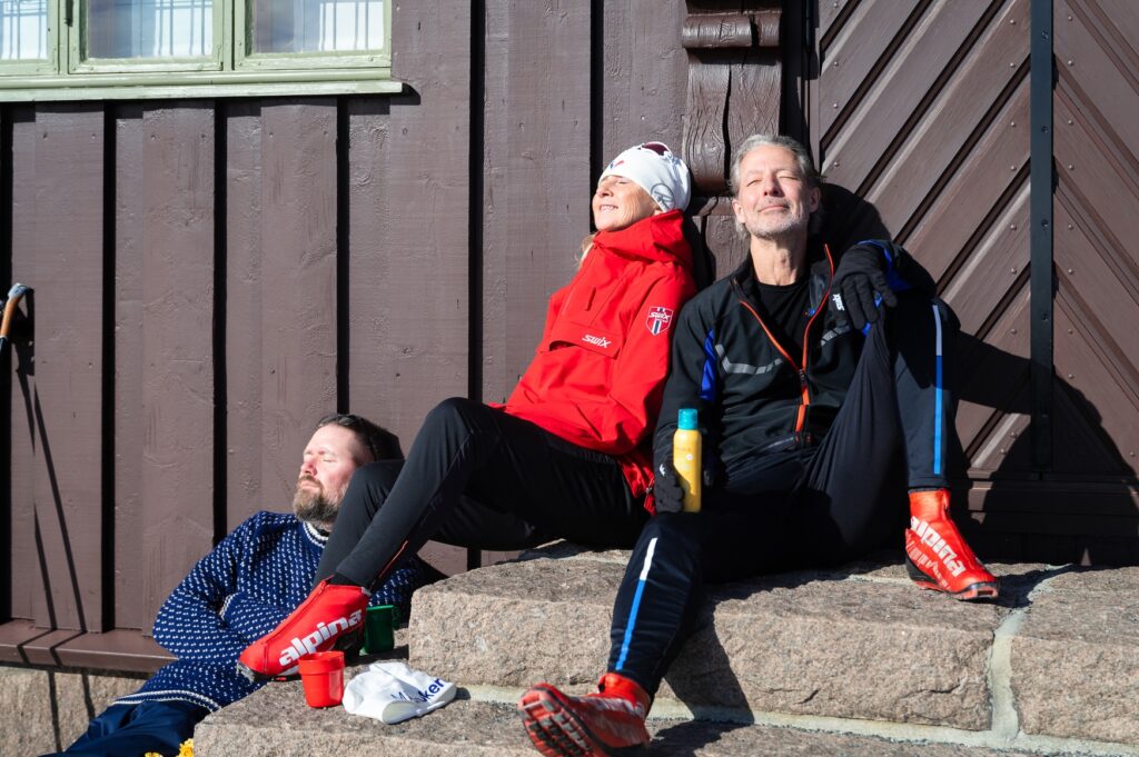 En kvinne og to menn sitter i solveggen og nyter sola etter å ha vært på skitur.