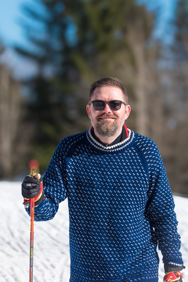 Mann som smiler mens han holder langrennsstaver, med snølandskap i bakgrunnen.