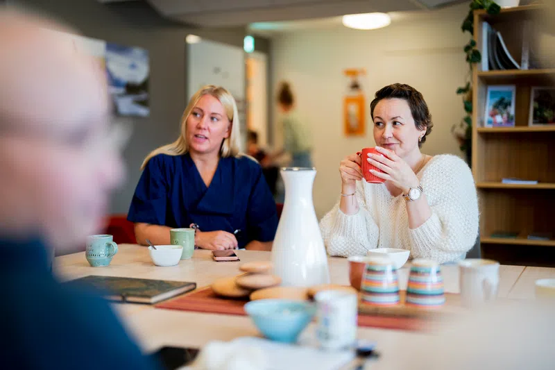 Folk sitter rundt et bord, en kvinne holder et krus