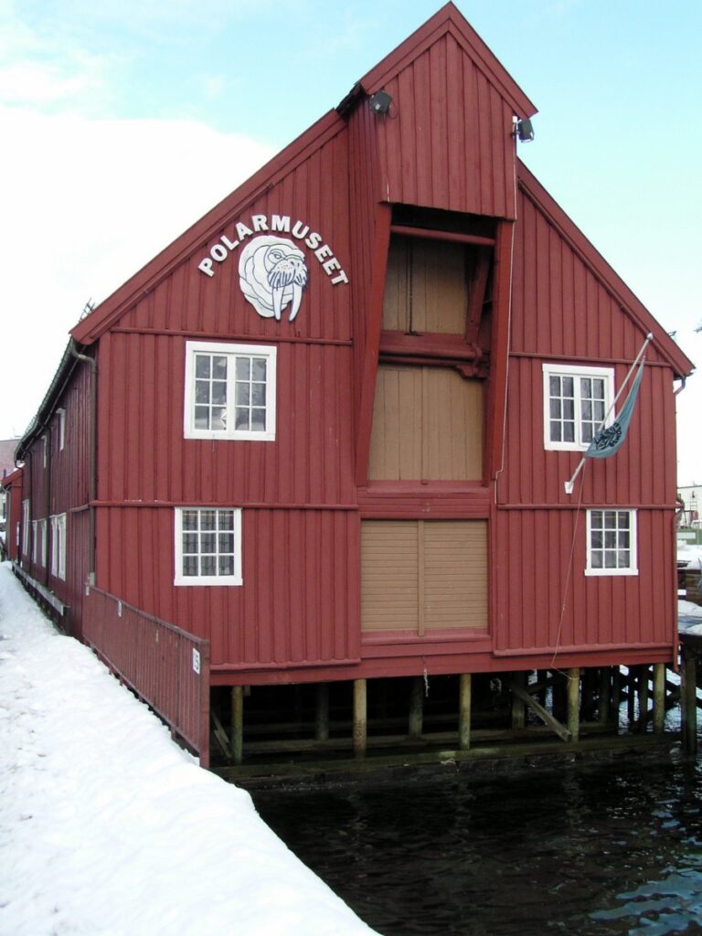 Rød trebygning på påler over snødekt mark, merket "polarmusee" med et isbjørnemblem, som indikerer at det er et ismuseum.