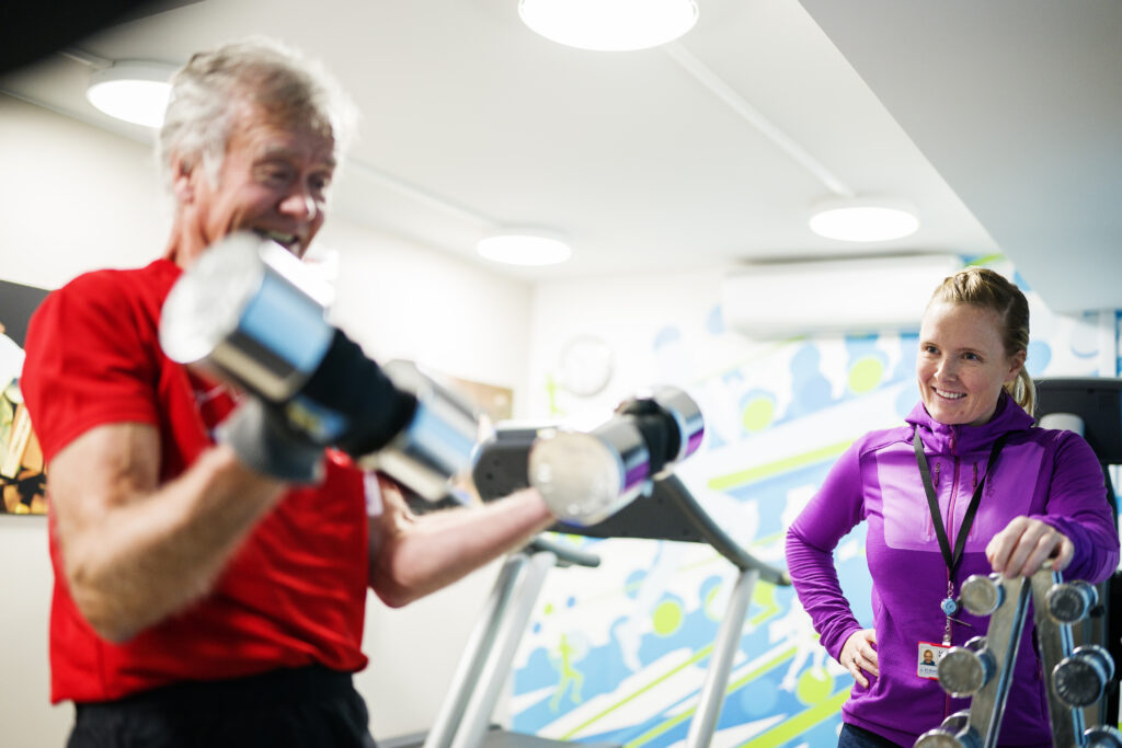 Senior mann som løfter manualer med hjelp fra en kvinnelig trener i et treningsstudio.