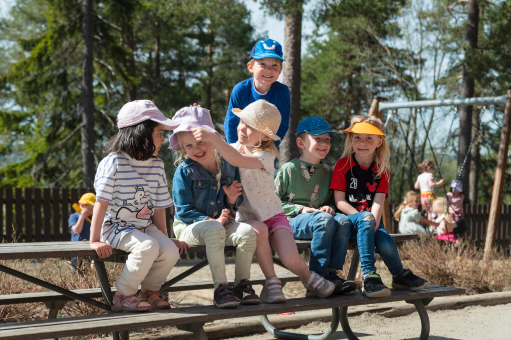 Seks barn iført fargerike hatter og sitter på en parkbenk, smiler og samhandler lekende på en solrik dag, med trær i bakgrunnen.