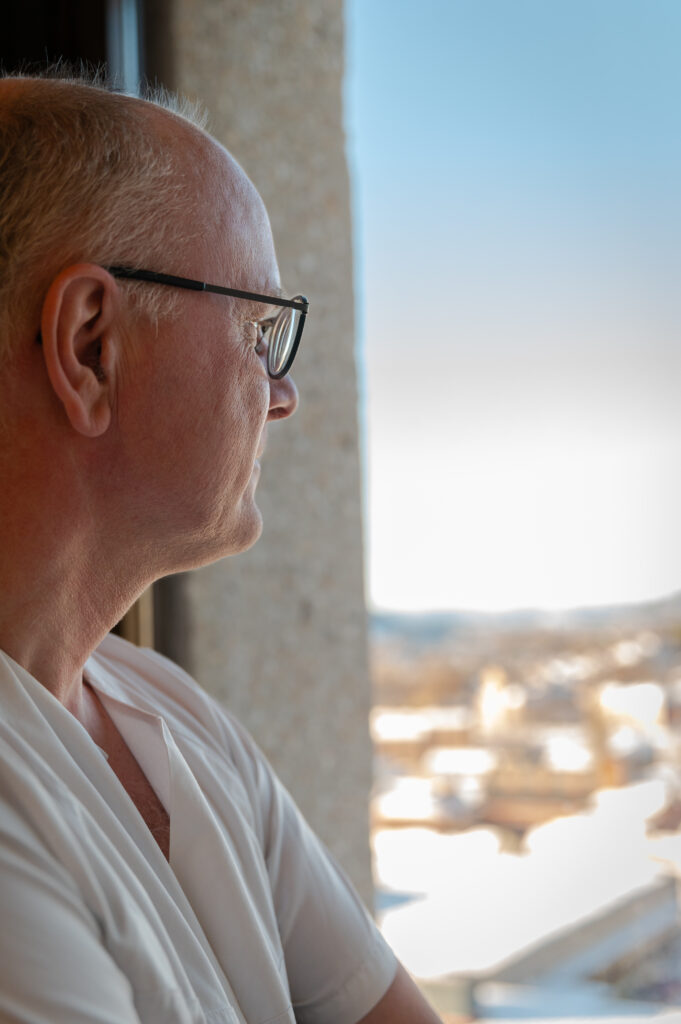 Middelaldrende mann med briller ser ut et vindu med et lyst, solrikt landskap synlig i bakgrunnen.