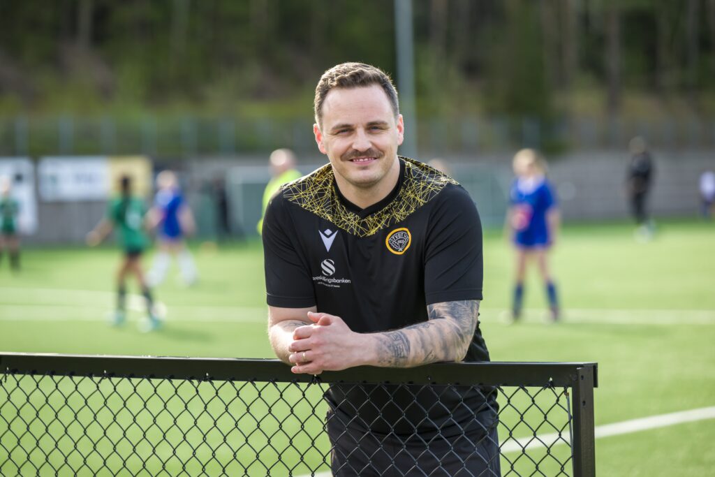 En mann i en svart sportsskjorte med gule detaljer lener seg på et gjerde med kjetting og smiler. En fotballkamp finner sted på banen bak ham.