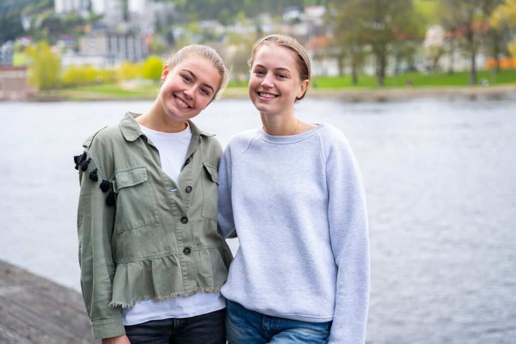 To jenter står ved en elv, holder rundt hverandre og smiler inn i kamera.