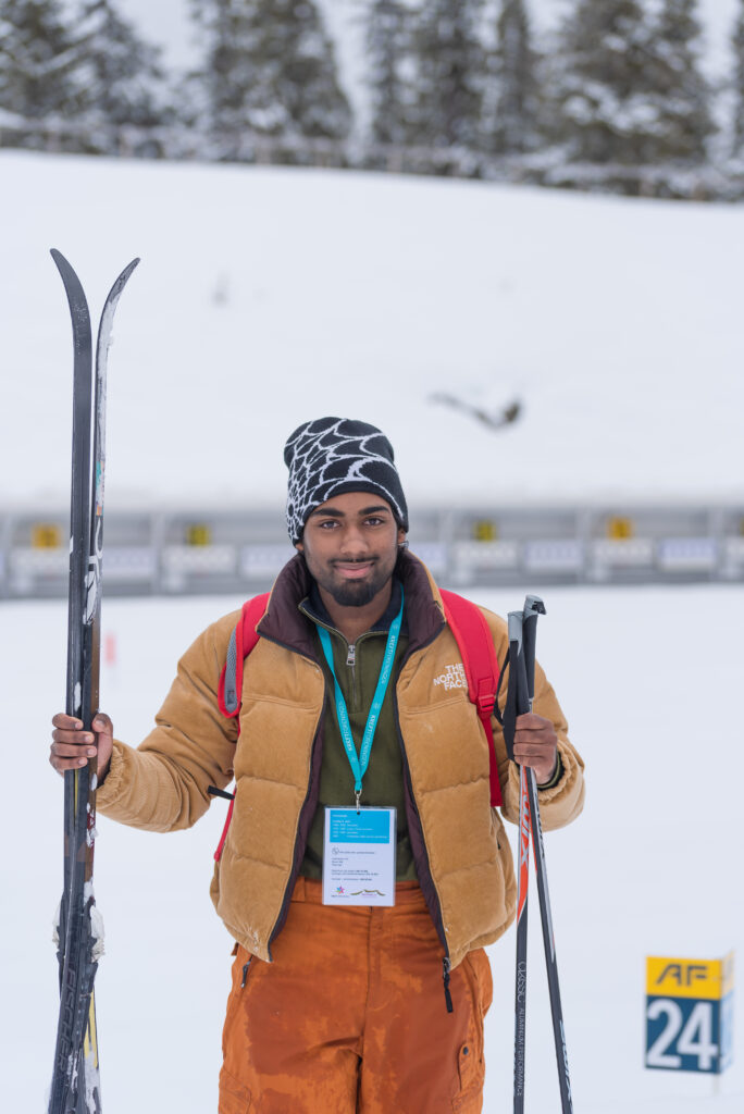 En person kledd i vinterklær med ski og skistaver står på snødekt mark med trær og en skiløype i bakgrunnen.