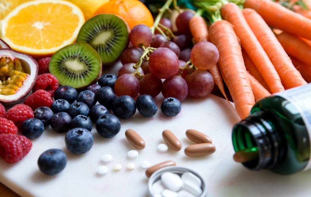 Assortert frukt og grønnsaker inkludert kiwi, appelsiner, druer, gulrøtter, blåbær og bringebær, vist med ulike piller og en grønn flaske på en hvit overflate.