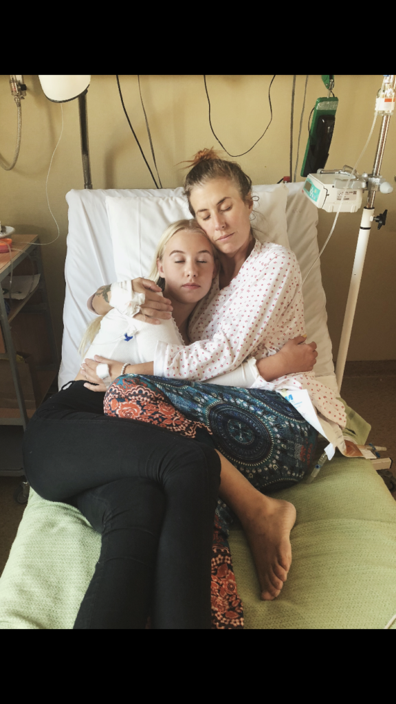 To kvinner, en med medisinsk bandasje på hånden, ligger sammen på en sykehusseng. De omfavner med lukkede øyne. Medisinsk utstyr er synlig i bakgrunnen.