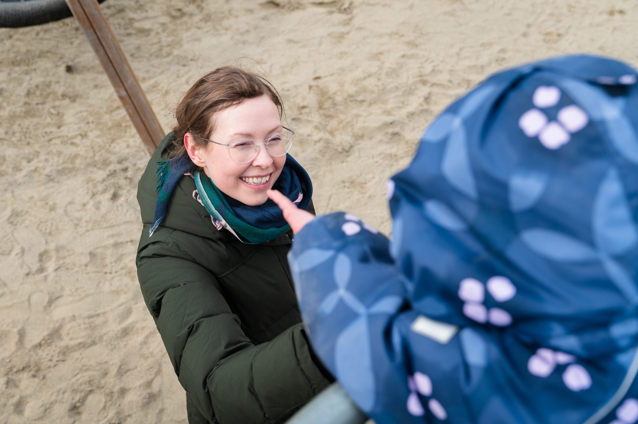 En kvinne med briller og grønn jakke smiler til en person i blåmønstret jakke på en sandstrand.