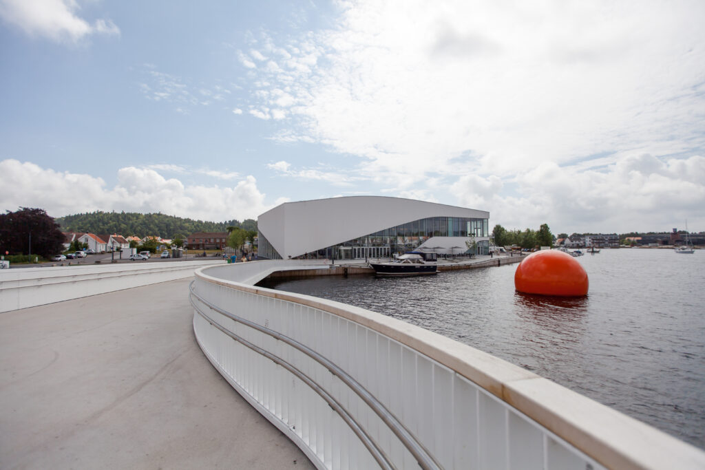 Moderne bygning ved vannkanten med buet arkitektur ved siden av en elv; en lys oransje bøye flyter i vannet, og skyer sprer seg over den blå himmelen over synlige betongstier og grønne omgivelser.