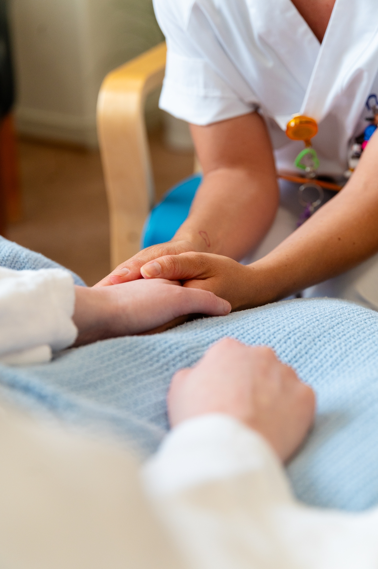 Sykepleier i hvit uniform holder forsiktig hånden til en pasient som ligger i en seng dekket med et lyseblått teppe.