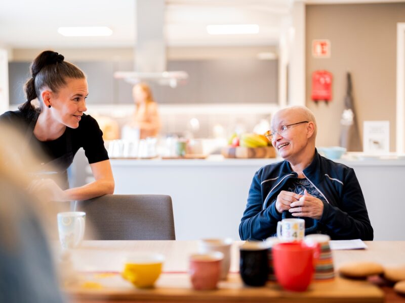 To personer snakker sammen i et lyst, moderne kjøkken. Den ene står og smiler, mens den andre, som sitter, lytter oppmerksomt. Ulike krus og snacks står på bordet i forgrunnen.