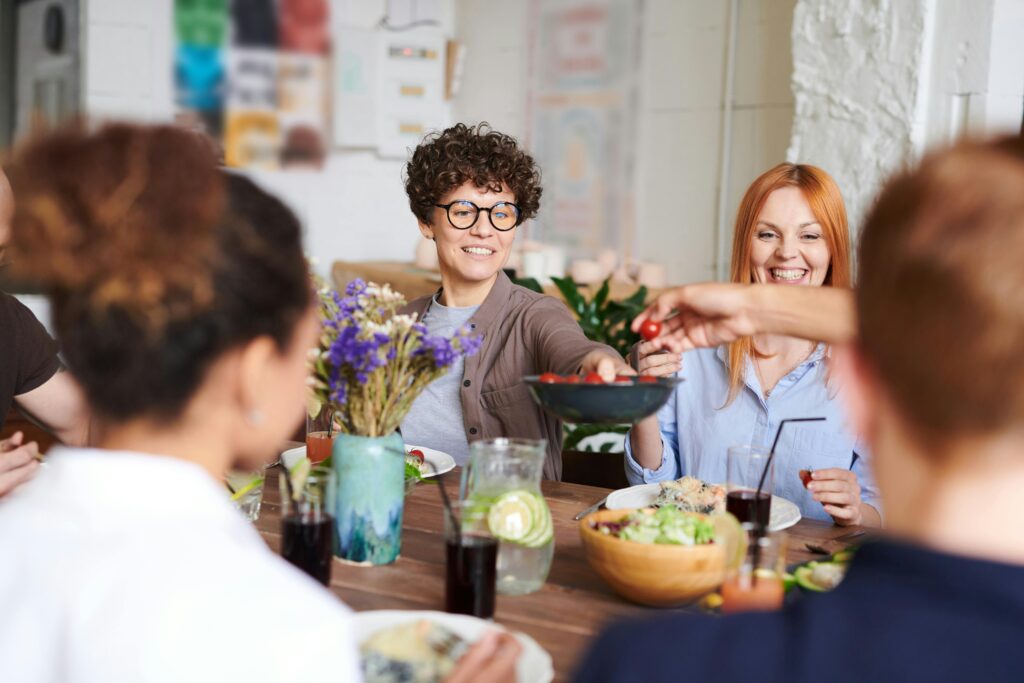 En gruppe mennesker sitter rundt et bord og nyter et måltid sammen. En person passerer en skål med mat mens andre smiler. En vase med blomster og drikke står også på bordet.