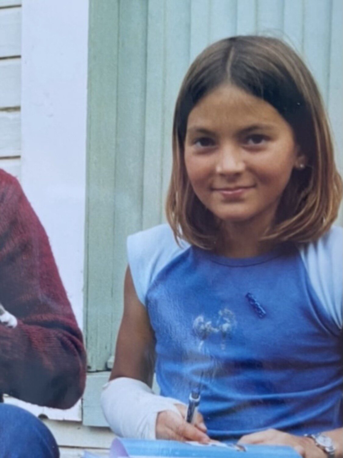 En ung jente med skulderlangt hår, iført en blå ermeløs skjorte, sitter utenfor ved et hus og holder en penn og notatbok. Hun har en bandasjert venstre underarm og smiler lett til kameraet.