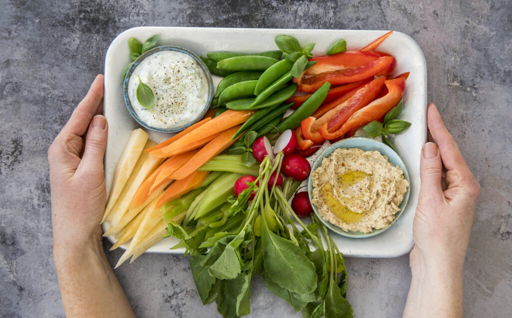 En persons hender som holder et brett med ferske grønnsaker inkludert gulrøtter, reddiker, paprika og selleri med boller med dip og hummus.