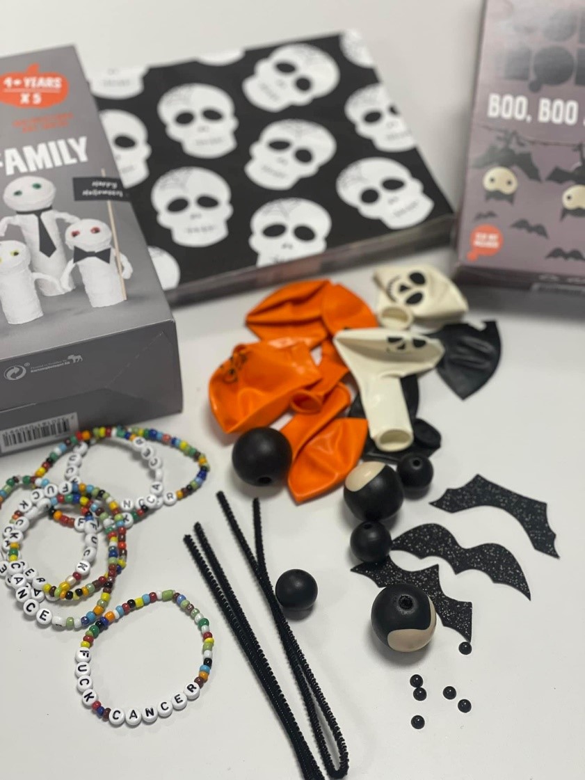 Diverse håndverksartikler med Halloween-tema, inkludert armbånd med perler, oransje og svarte ballonger, piperensere og små dekorative gjenstander er arrangert på en hvit overflate.