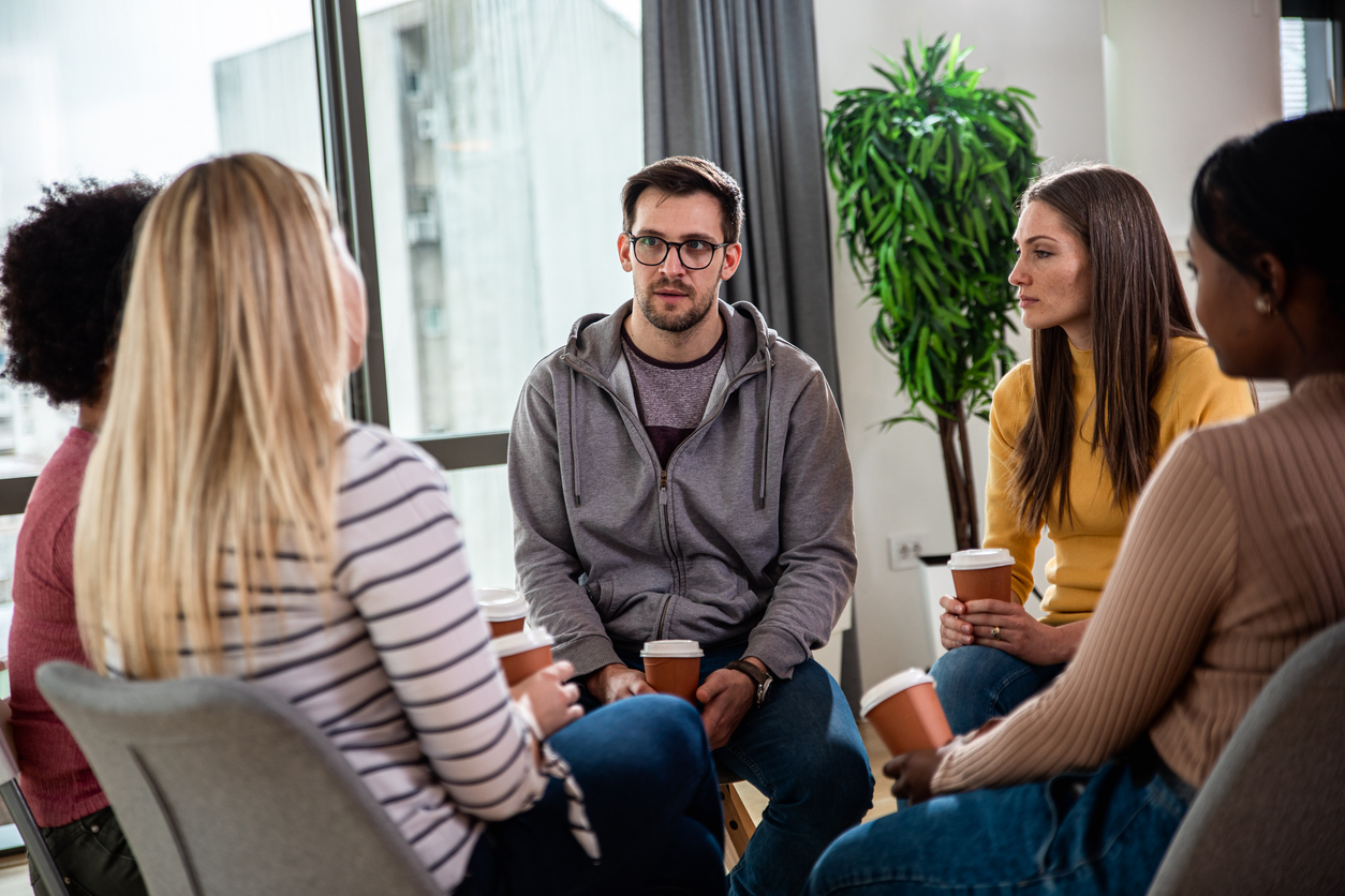 En gruppe på fem personer sitter i ring, snakker og holder kaffekopper. Det er en plante og et vindu i bakgrunnen.