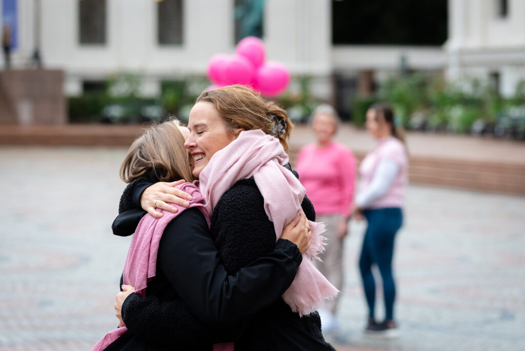 To personer klemmer mens to andre står i bakgrunnen. Gruppen er utendørs, med rosa ballonger synlige bak.