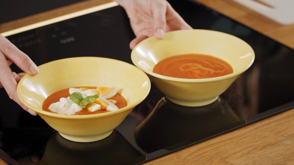 To gule boller med tomatsuppe på en benkeplate. Den ene bollen er vanlig mens den andre er pyntet med hakkede egg, rømme og et basilikumblad. En persons hender holder bollene.