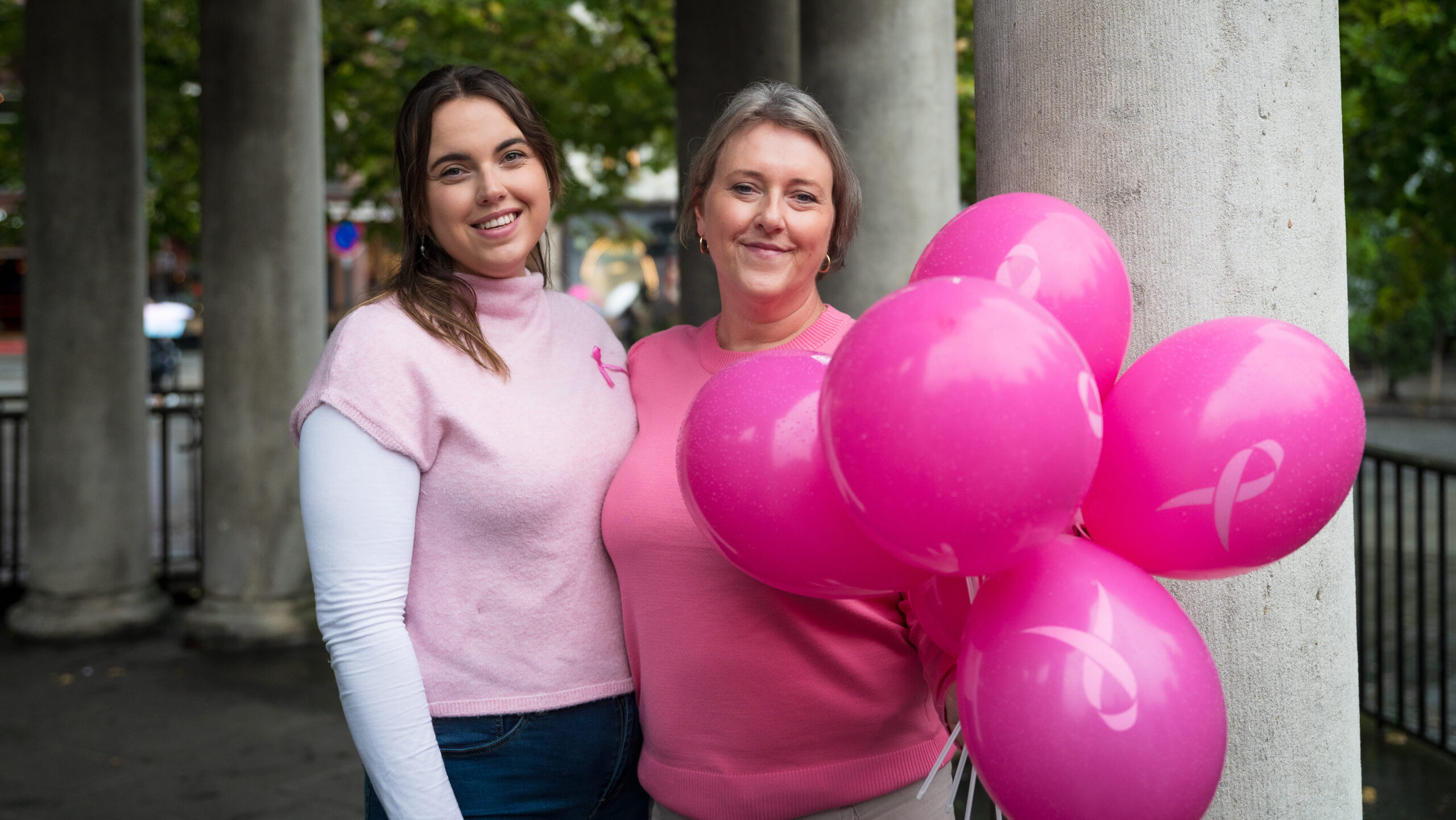 To kvinne rikledd rosa holder rundt hverandre og ser i kamera, begge smiler. Ene kvinnen holder rosa ballonger. 