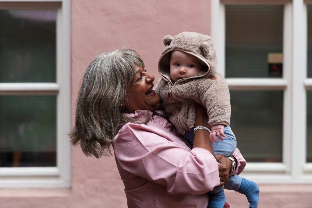 En kvinne smiler og holder barnebarnet sitt. Babyen har på en brun teddygenser med ører. 