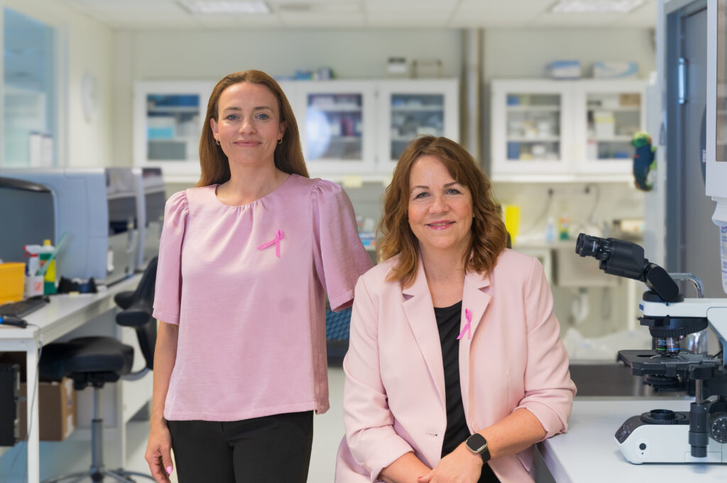 To kvinner i rosa antrekk med rosa bånd er i et laboratorium. Den ene står mens den andre sitter i nærheten av et mikroskop. Laboratoriet har diverse utstyr og hyller i bakgrunnen.