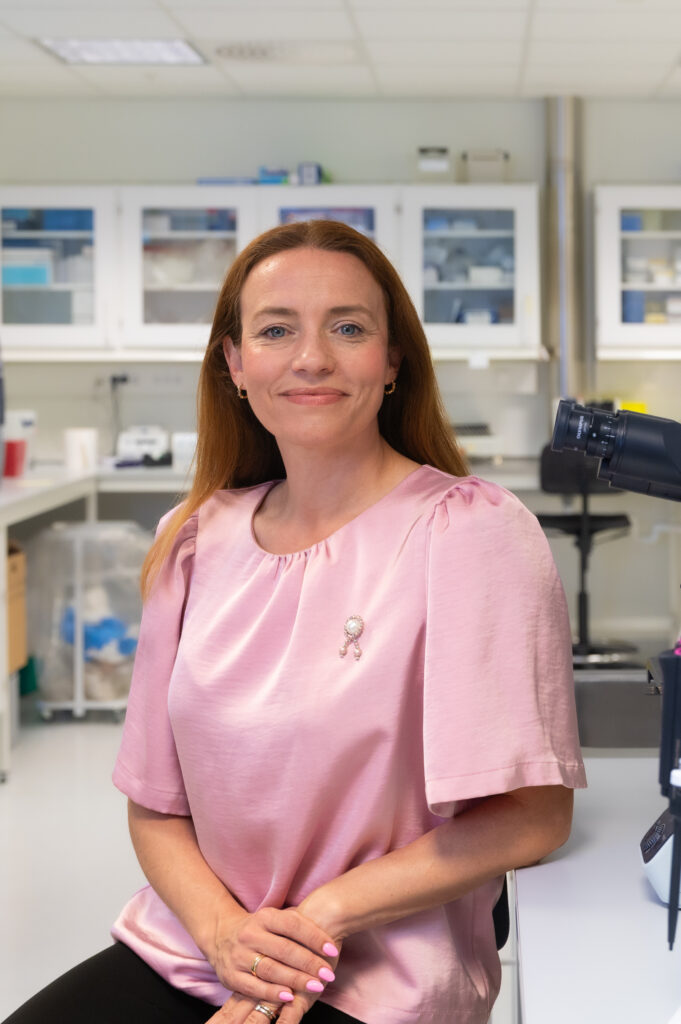 En person med langt hår, iført en rosa bluse og med årets Rosa sløyfe på, sitter i en laboratoriesetting med hyller og utstyr synlig i bakgrunnen.