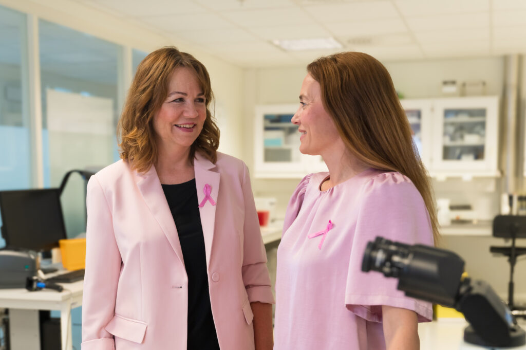 To kvinner iført rosa klær med rosa silkesløyfe på og snakker sammen i laboratoriemiljø.