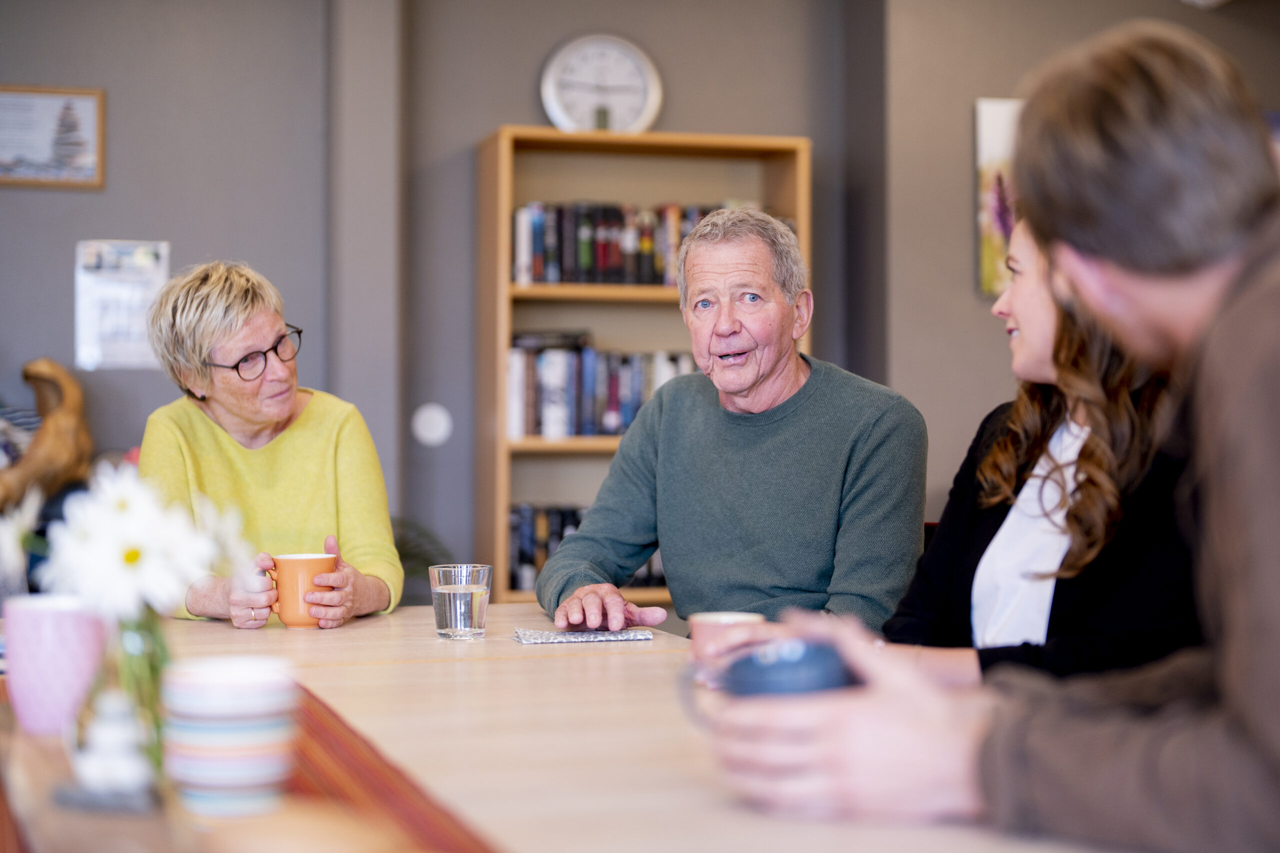 En gruppe på fire personer, inkludert en eldre mann og kvinne, sitter rundt et bord i uformelle omgivelser, i samtale. Bokhyller og en klokke er synlige i bakgrunnen.