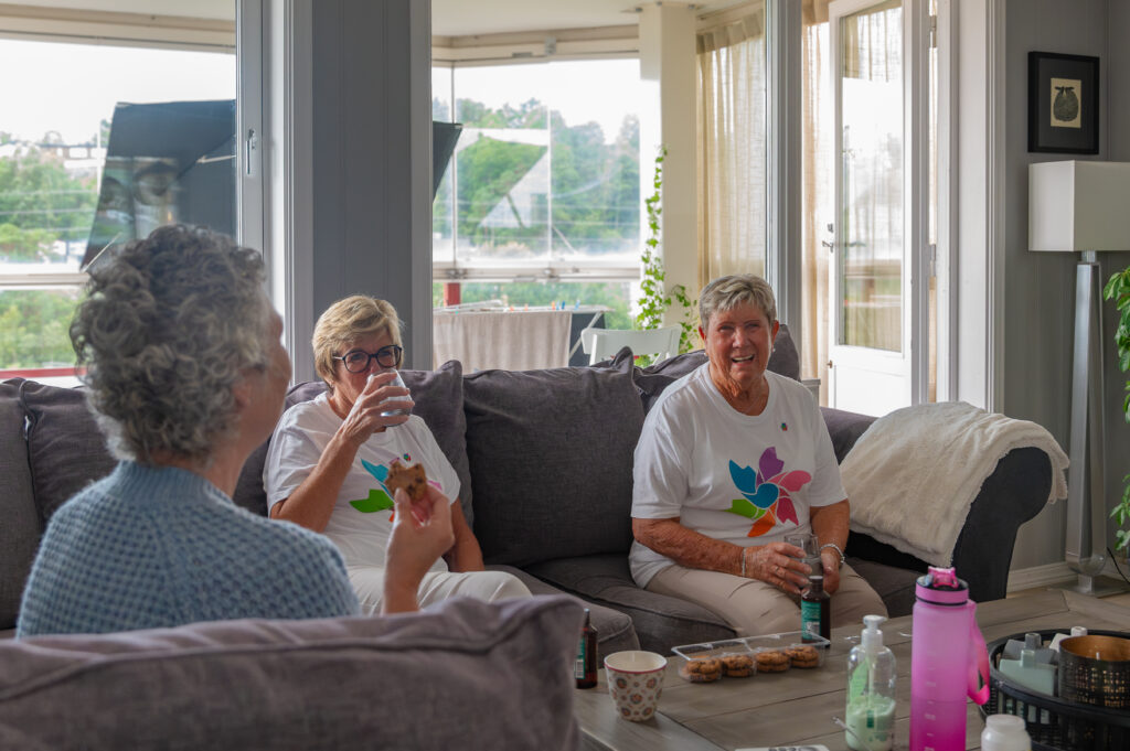 Tre kvinner som sitter på en sofa i en stue, prater og holder drinker. Snacks og drikke står på bordet foran dem. Det kommer sterkt dagslys gjennom vinduene.