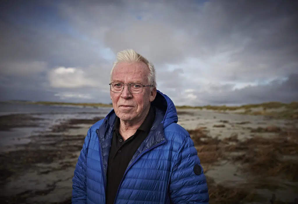 En eldre mann i blå jakke står på en strand, med overskyet himmel og hav i bakgrunnen.