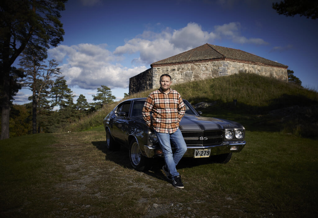 En person i rutete skjorte står ved siden av en klassisk svart bil parkert på et gresskledd område foran en historisk steinbygning under en delvis overskyet himmel.